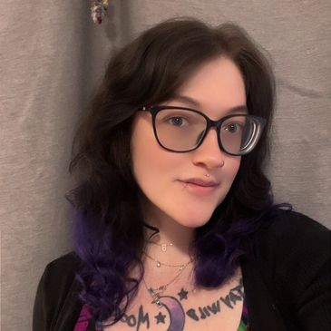 A young woman with glasses and purple-tinted hair poses indoors.