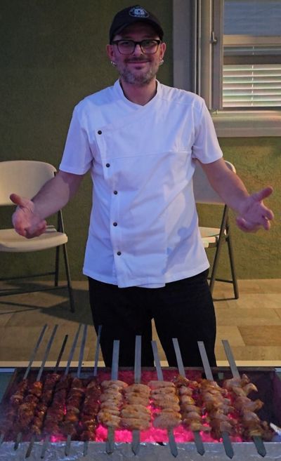 Chef grilling skewers of meat over glowing coals outdoors.