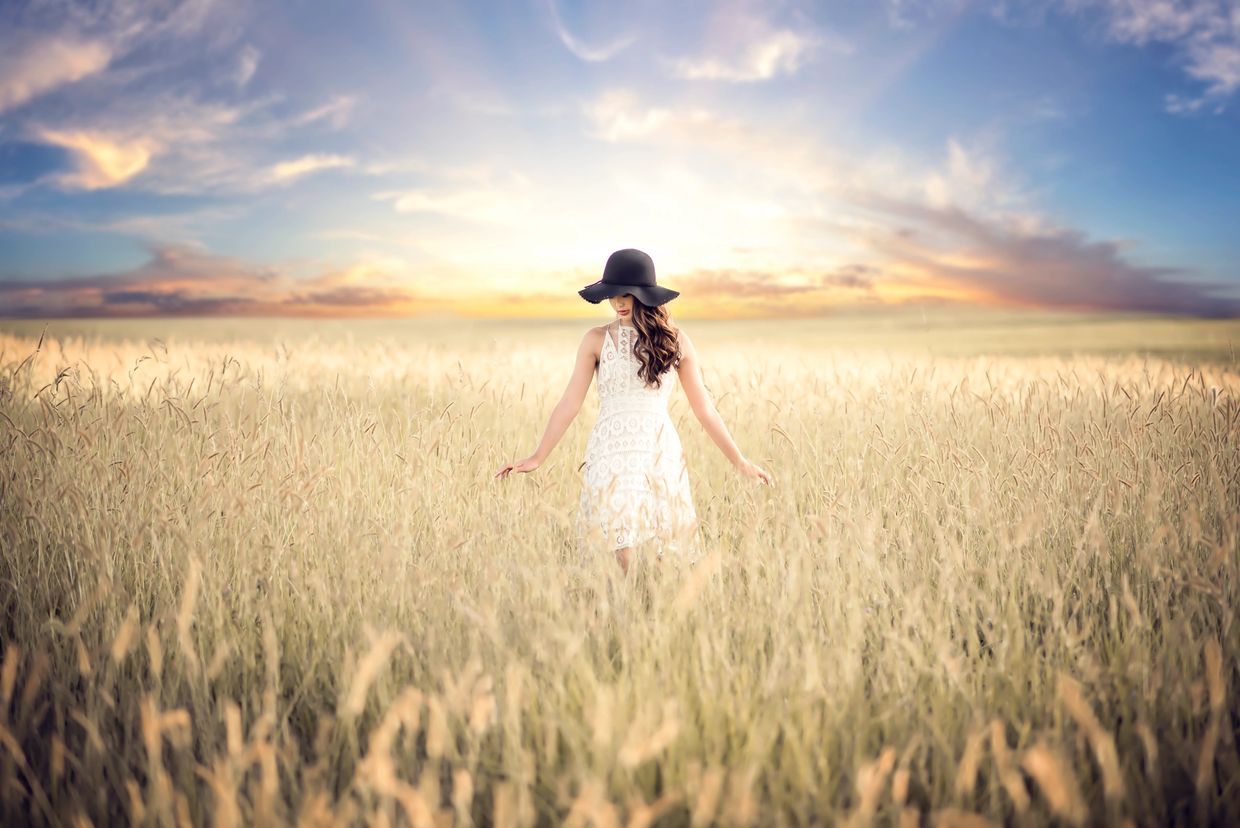 Woman in white dress and black hat walking through a wheat field at sunset.