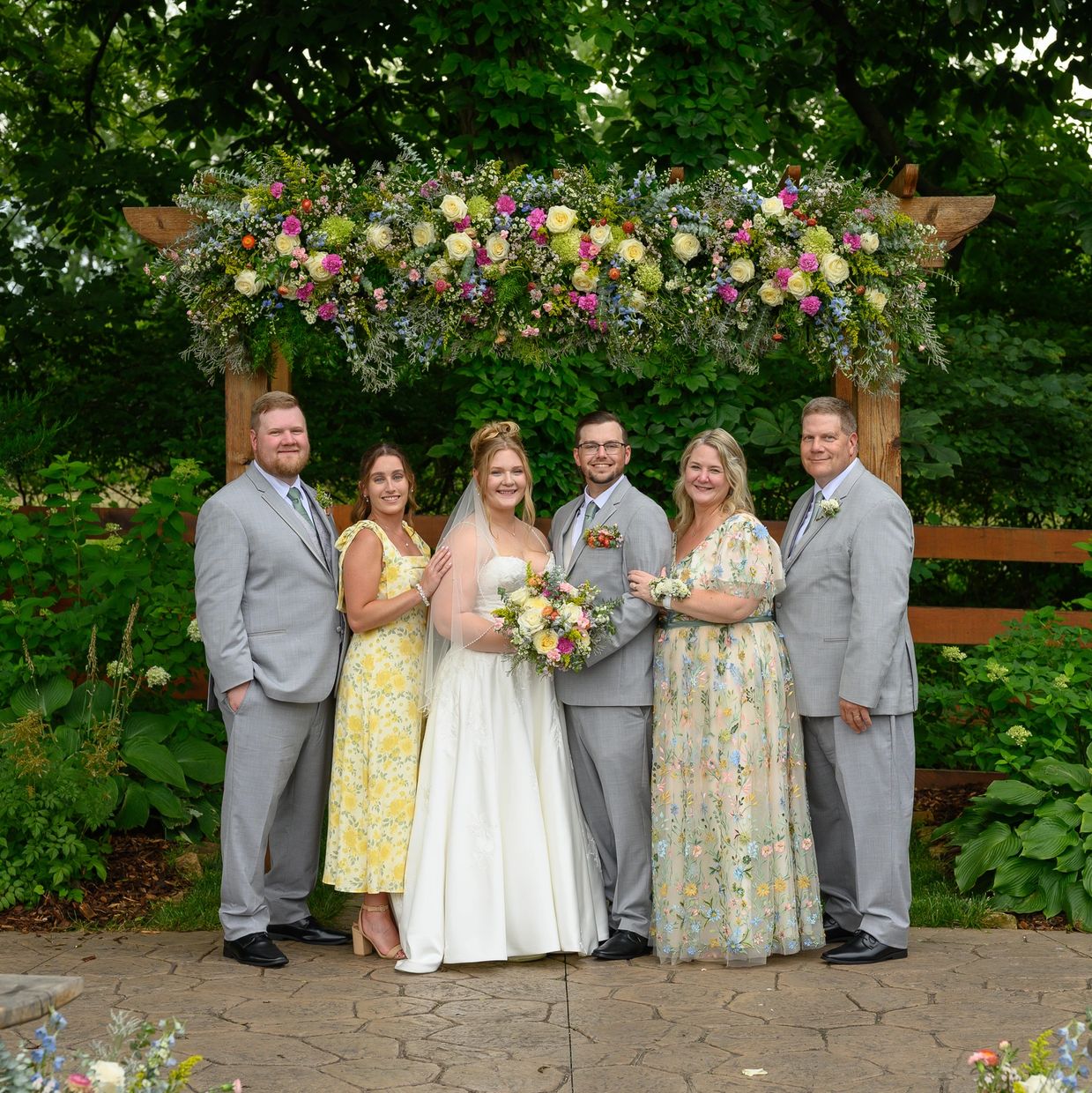 Wedding party under floral arch outdoors.