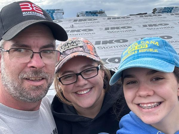 Three people smiling wearing caps, standing outdoors near a roof under construction.