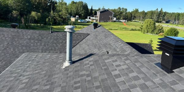 View of a gray shingled roof with chimney vents and a green landscape background.