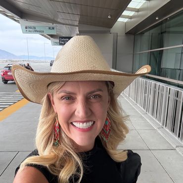 Smiling woman wearing a large straw hat and colorful earrings at an airport.