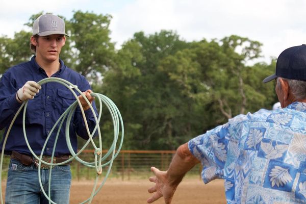 A young man prepares a lasso while an older man gestures towards him outdoors.