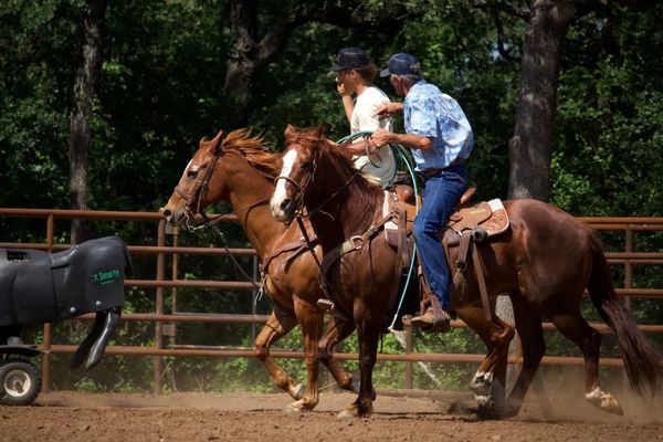 Two men riding horses in an outdoor arena with a mechanical bull nearby.