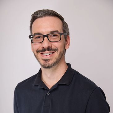 Smiling man with glasses in a black polo shirt.