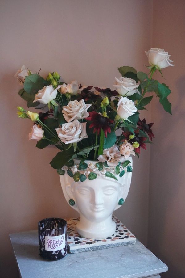 White and dark flowers in a decorative head-shaped vase on a table with a candle.