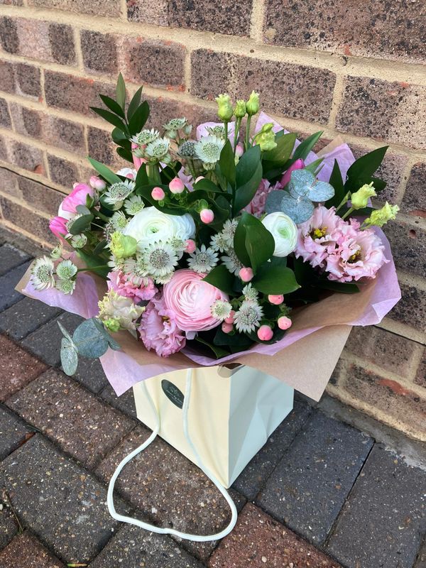 A pastel flower bouquet in a cream gift bag outside against a brick wall.