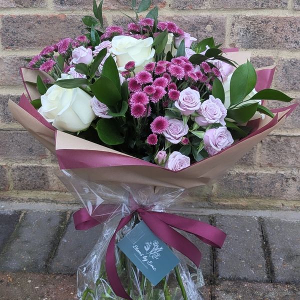 A beautiful bouquet of white roses, pink chrysanthemums, and lavender roses wrapped in brown and purple paper.