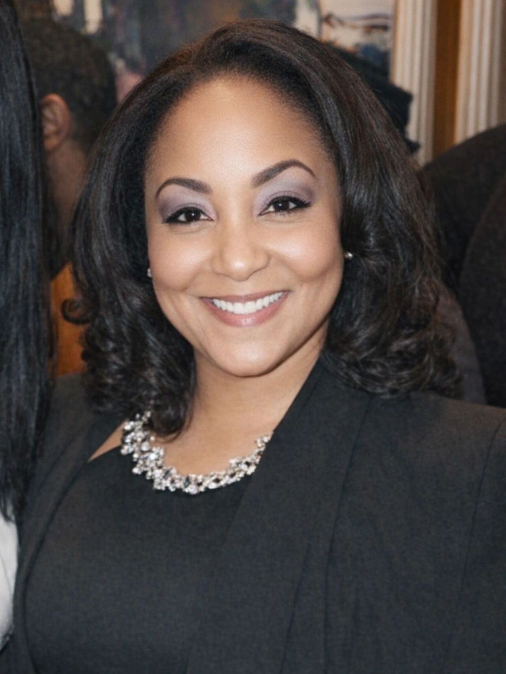Smiling woman with curly hair wearing a black outfit and a sparkling necklace.