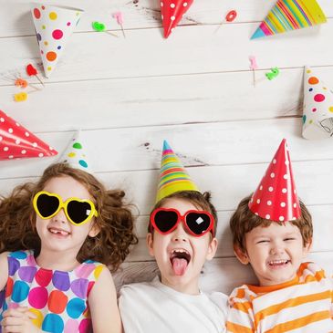 Three kids in party hats and heart-shaped sunglasses lying on the floor, smiling and having fun.