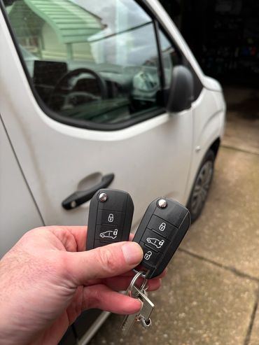 Hand holding two car key fobs in front of a white vehicle.