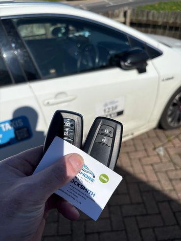 Hand holding two car keys and a locksmith card in front of a white car.