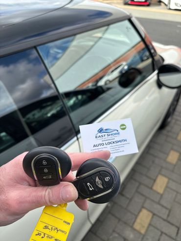 Hand holding two car keys and an auto locksmith card near a white car.