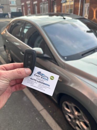 Hand holding car key and locksmith business card near a parked car.