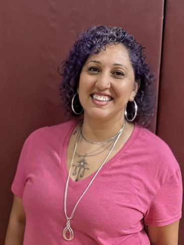 Smiling woman with curly purple hair wearing a pink shirt and silver jewelry.