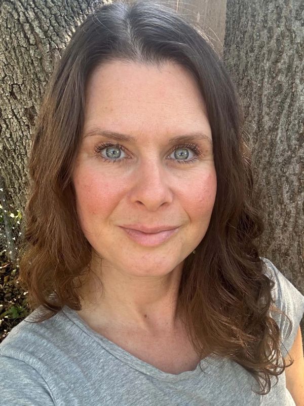 Woman with blue eyes smiling outdoors near a tree.