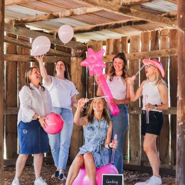 Women celebrating with pink balloons and a magic wand in a rustic wooden barn.