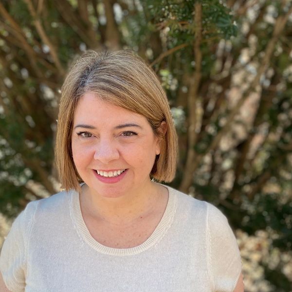Smiling woman with short blonde hair standing outdoors in front of trees.