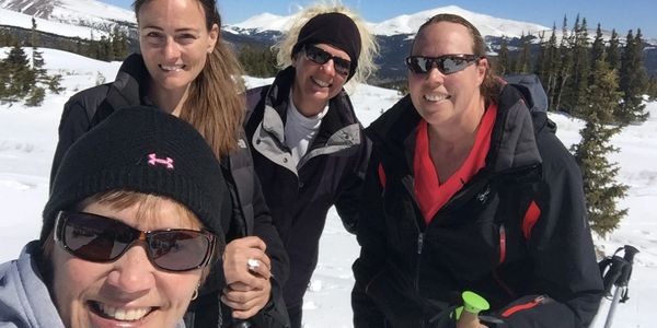 Four women smiling outdoors in snowy mountains.