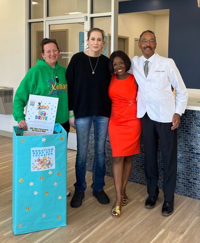 Four people stand posing by a Kellan's Legacy of Love toy drive donation box in a dentist office.
