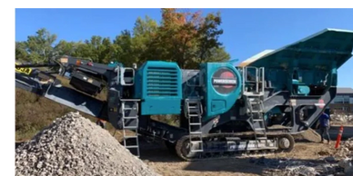 Large blue stone crusher machine at a construction site with gravel piles.