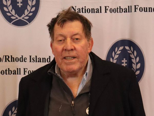 An elderly man in a dark jacket stands in front of a football foundation backdrop.