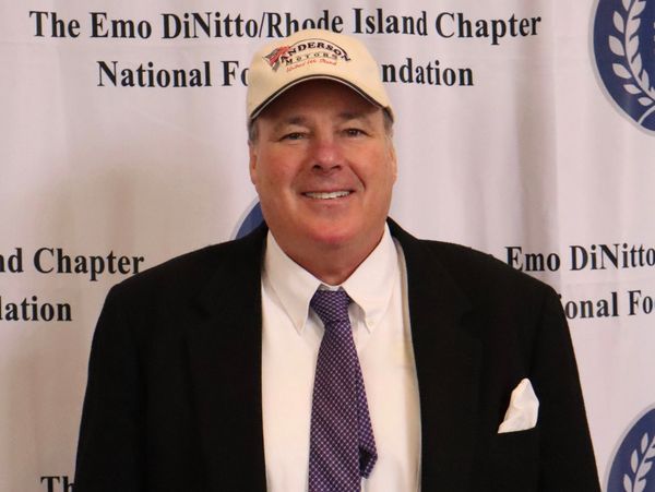 Man in a suit and tie wearing a baseball cap at a formal event backdrop.