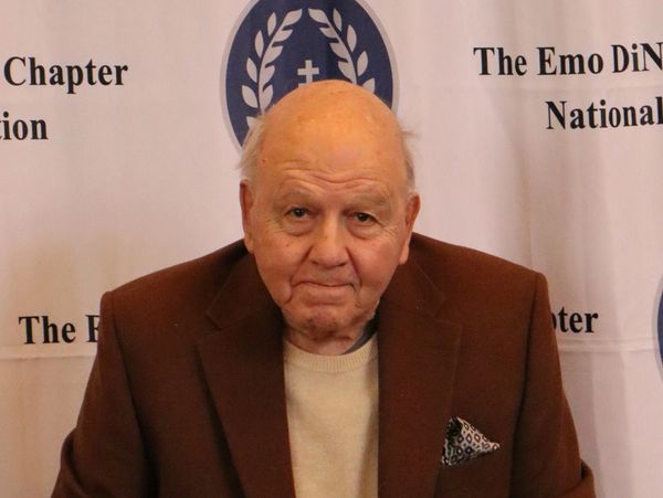 Elderly man in a brown jacket posing at an event backdrop.