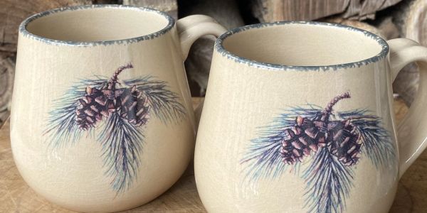 Two cream-colored mugs with pinecone and pine needle designs.