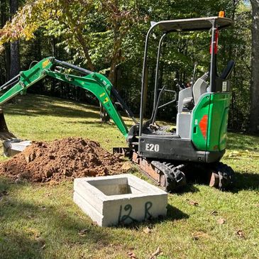 Mini excavator digging near a concrete box labeled '12R' in a grassy area.