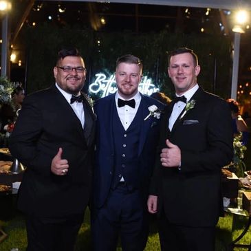Three men in tuxedos posing happily at a nighttime event.