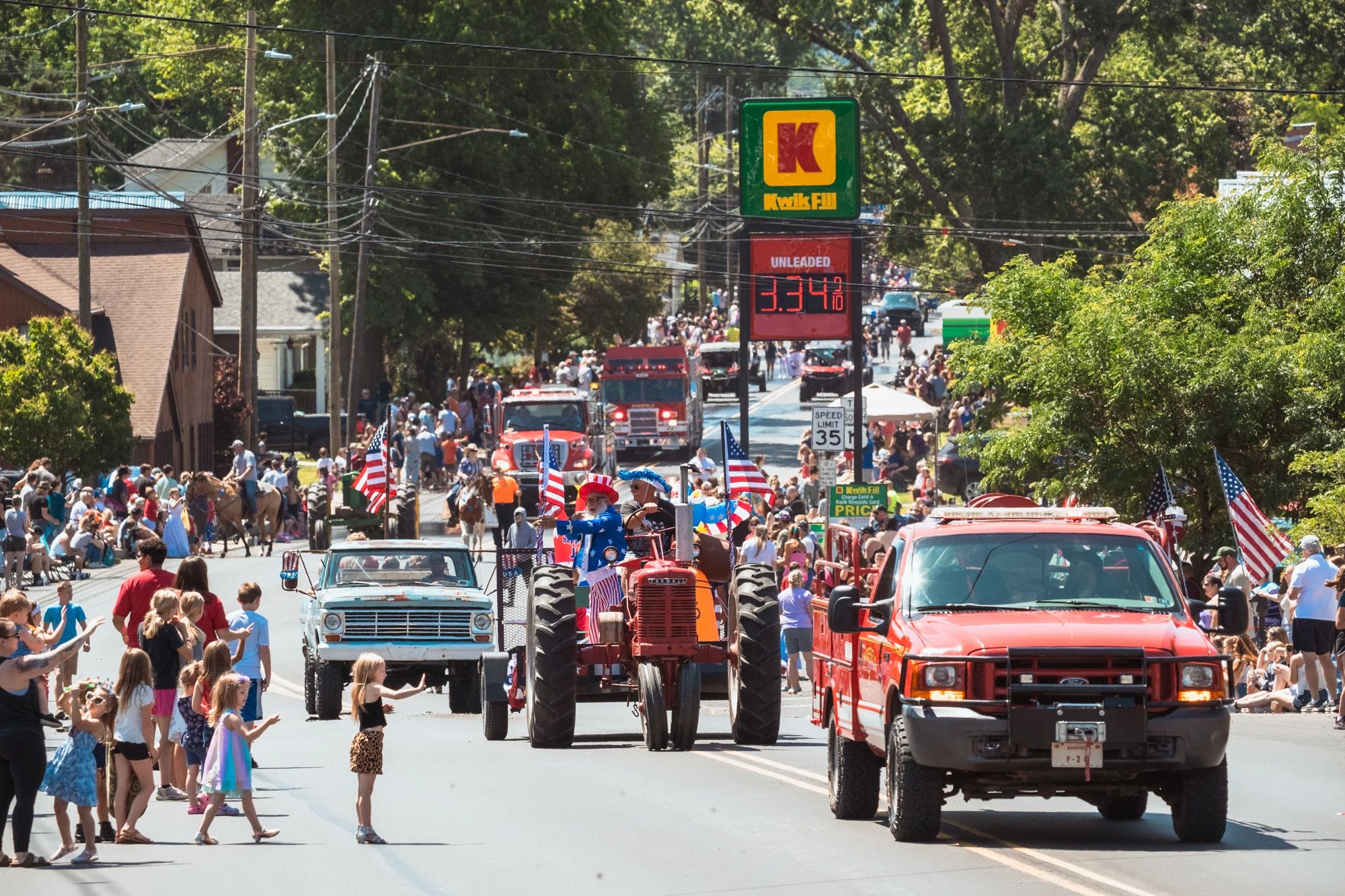 A patriotic parade with people, tractors, and vehicles on a sunny day.