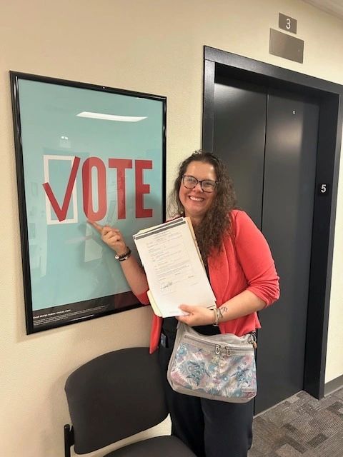 A woman smiling and pointing to a vote poster while holding documents.