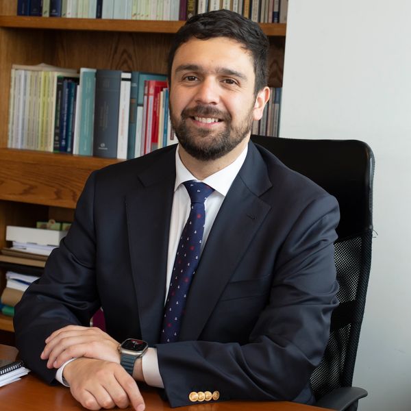 Confident man in a suit sitting at a desk with books in the background.