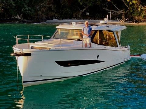 A man stands on a sleek white motorboat in clear turquoise water near a leafy shoreline.