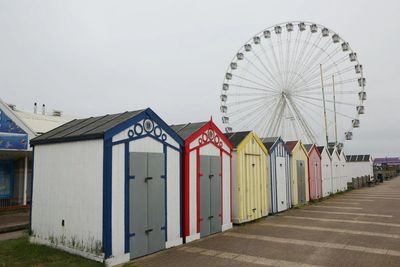 Colorful beach huts lined up with a Ferris wheel in the background on a cloudy day.