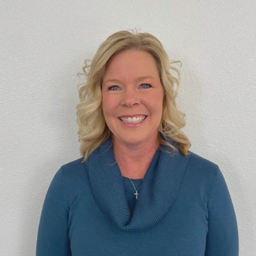 Smiling woman in a blue sweater with a cross necklace against a white wall.