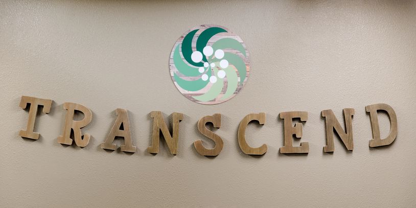 Wooden letters spelling TRANSCEND on a wall with a circular logo above.