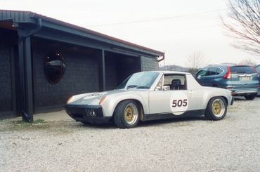 Silver vintage race car with number 505 parked on gravel near a building.