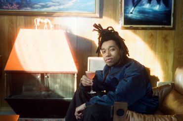 A young man with dreadlocks holding a cocktail in a cozy, wood-paneled room.