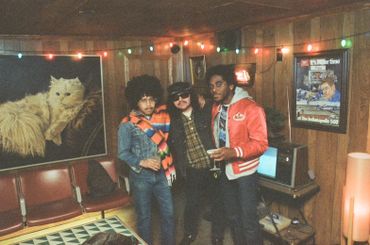 Three men posing in a retro room with wood paneling and colorful lights.