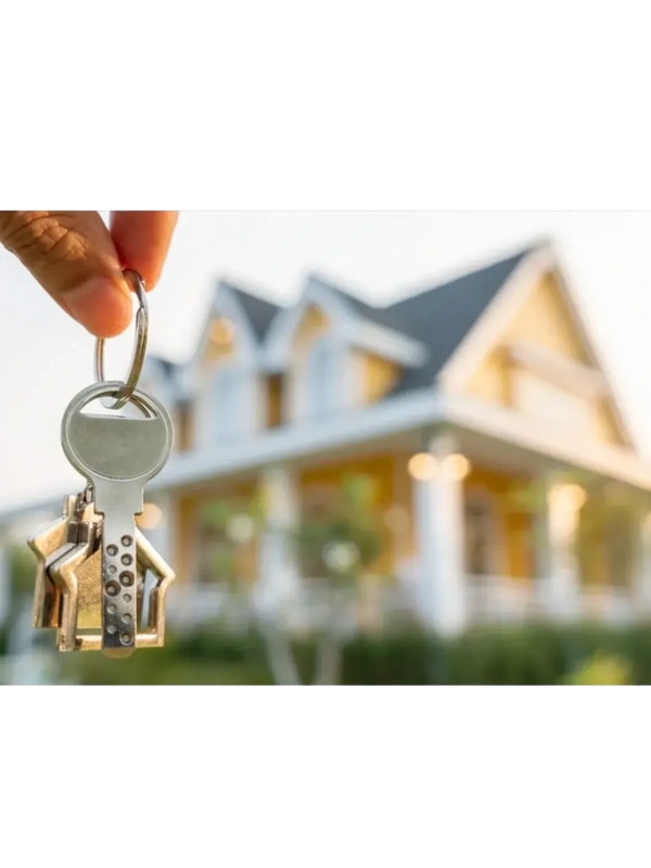Hand holding house-shaped keys in front of a blurred house.