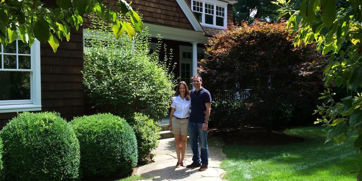 Smiling couple standing proudly in front of a luxurious new home with lush plantings around it