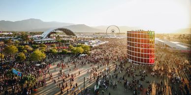 Crowded outdoor festival with stage, ferris wheel, and sunset.