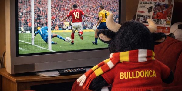 A bull mascot watches a soccer game on TV wearing a scarf and jersey.