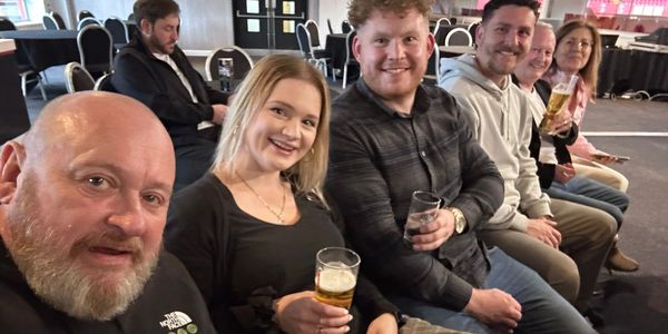 A group of six people sitting indoors, holding drinks and smiling at the camera.