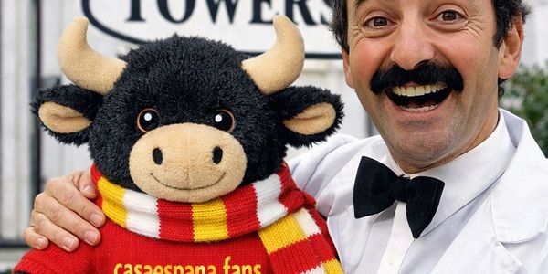 A man in a white suit and bow tie smiles next to a stuffed bull wearing a red scarf.