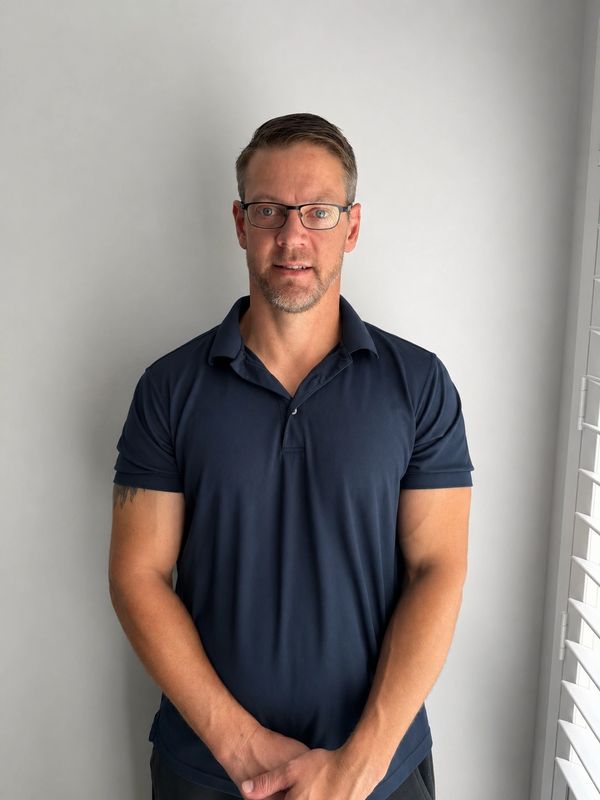 Man wearing glasses and a navy blue polo shirt standing against a white wall.