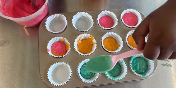 Hand filling cupcake liners with colorful batter using a pink spoon.